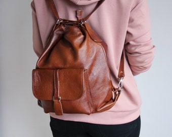 May include: A brown leather backpack with a drawstring closure and a front pocket. The backpack is being worn by a person wearing a pink hoodie.