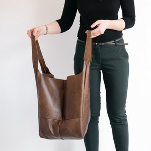 May include: A large, unstructured brown leather tote bag with a smooth finish and two wide shoulder straps. The bag features a deep, open main compartment and a rectangular base, showcasing a minimalist design with visible stitching along the seams.
