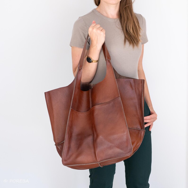 May include: A large brown leather tote bag with a single pocket on the front. The bag is being held by a person wearing a light brown t-shirt and dark green pants.