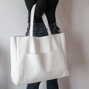 May include: A large, white leather tote bag with a front pocket and two handles. The bag has a rectangular shape and is made of a textured material. The handles are attached to the top of the bag with reinforced stitching. The bag is a neutral color.