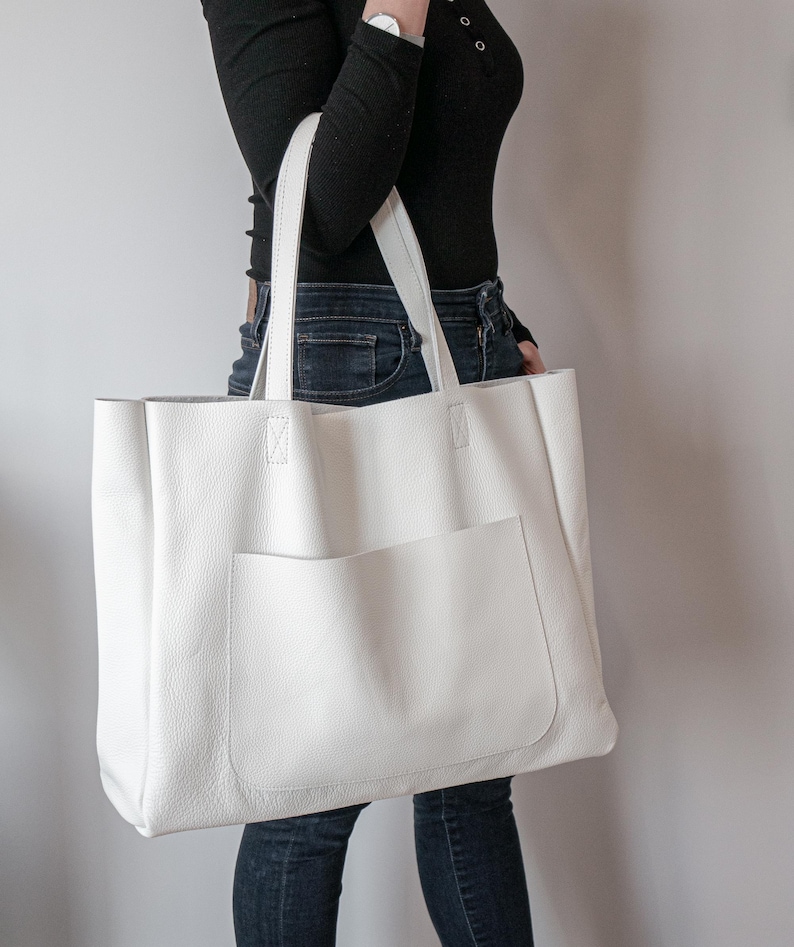 May include: A large, white leather tote bag with a front pocket and two handles. The bag has a rectangular shape and is made of a textured material. The handles are attached to the top of the bag with reinforced stitching. The bag is a neutral color.