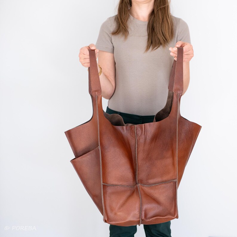 May include: A large brown leather tote bag with multiple pockets and a wide opening. The bag has long straps and is being held by a person.
