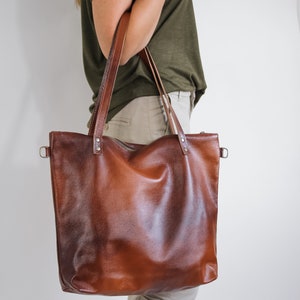 May include: A large, rectangular brown leather tote bag with a textured surface, featuring two long shoulder straps attached with silver-toned metal rivets, a top zipper closure, and side metal D-rings for additional strap attachments.