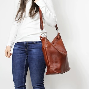 May include: A brown leather shoulder bag with a long strap. The bag is worn by a person wearing a white long-sleeved shirt and blue jeans.