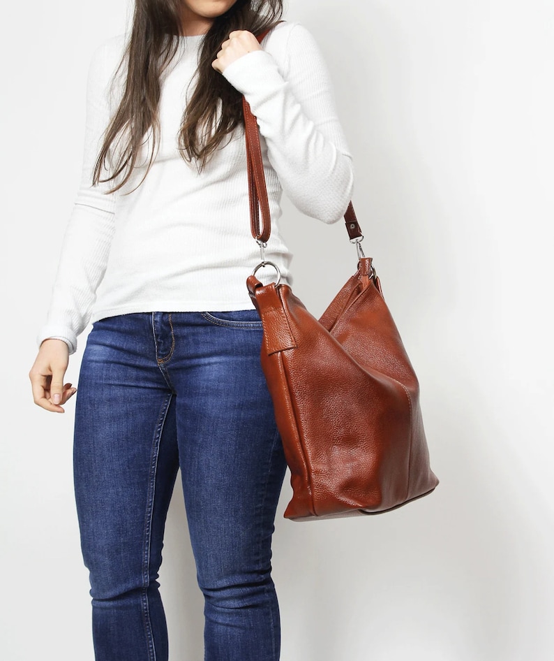 May include: A brown leather shoulder bag with a long strap. The bag is worn by a person wearing a white long-sleeved shirt and blue jeans.