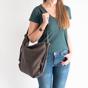 May include: A brown leather shoulder bag with a long strap. The bag is being worn by a person wearing a teal shirt and blue jeans.