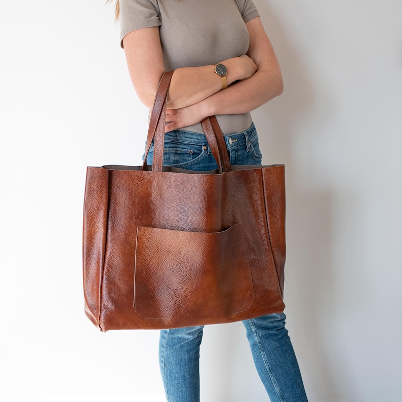 May include: A large brown leather tote bag with a front pocket. The bag has a wide opening and is made of a soft, supple leather.