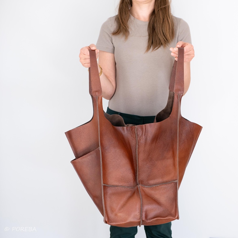 Puede incluir: Un bolso tote grande de cuero marr&oacute;n con varios bolsillos y una abertura ancha. El bolso tiene correas largas y lo est&aacute; sujetando una persona.