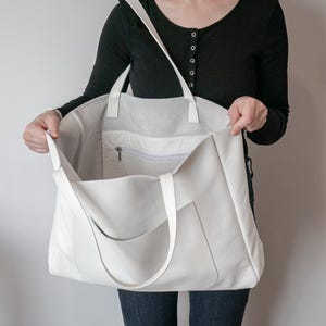 May include: A large, off-white leather tote bag with a shoulder strap and two handles. The bag has an exterior pocket and a zippered interior pocket. The bag is held open, revealing the spacious interior.