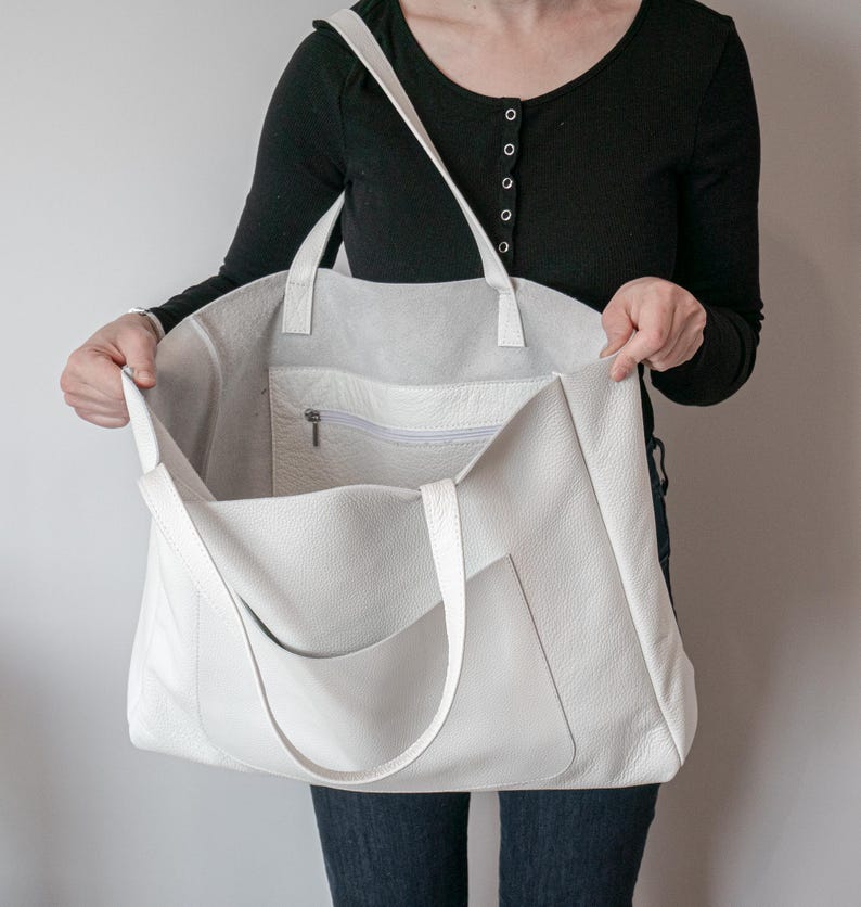 May include: A large, off-white leather tote bag with a shoulder strap and two handles. The bag has an exterior pocket and a zippered interior pocket. The bag is held open, revealing the spacious interior.