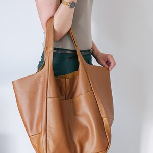 K&ouml;nnte beinhalten: Eine gro&szlig;e, weiche Tragetasche aus hellbraunem Leder mit einer glatten, genarbten Textur. Die Tasche verf&uuml;gt &uuml;ber zwei breite Schulterriemen, ein ger&auml;umiges Hauptfach und eine sichtbare Au&szlig;entasche auf der Vorderseite in einem weichen, formlosen Design.