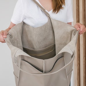 May include: A large, light grey leather tote bag with a zipped pocket on the inside. The bag is open and the lining is visible.