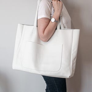 May include: A large, white tote bag with a rectangular front pocket. The bag has two long handles and is made of a smooth, leather-like material. The bag is being carried over the shoulder.