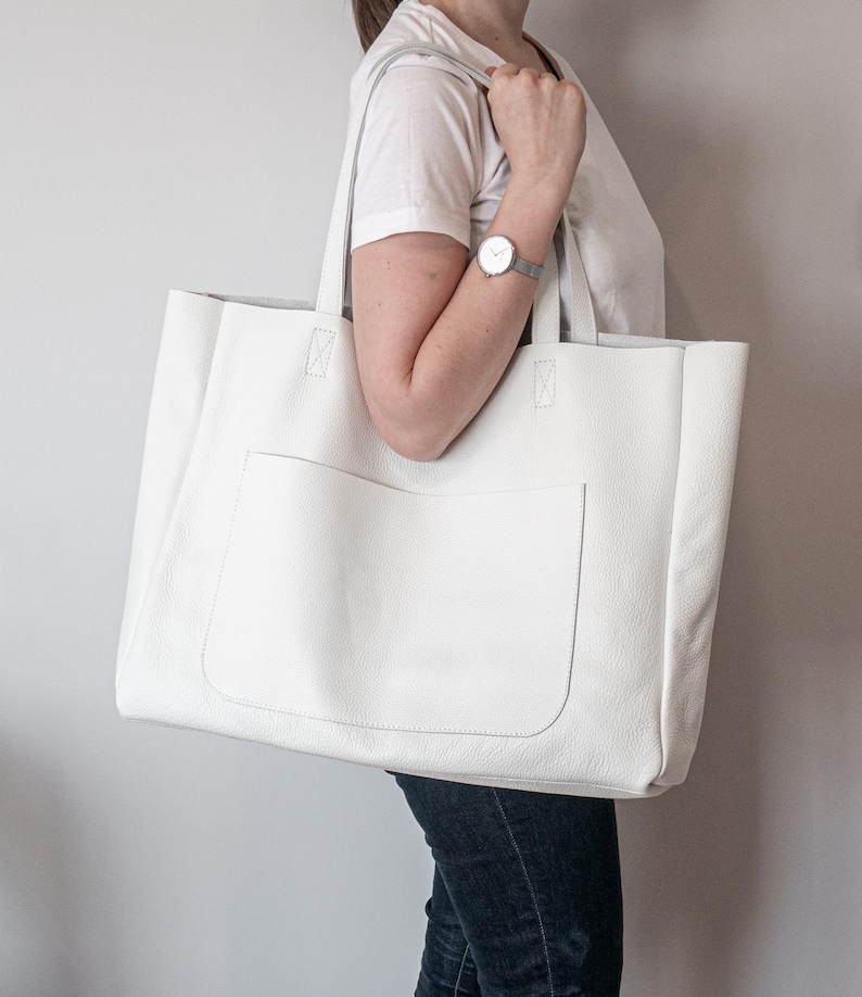 May include: A large, white tote bag with a rectangular front pocket. The bag has two long handles and is made of a smooth, leather-like material. The bag is being carried over the shoulder.