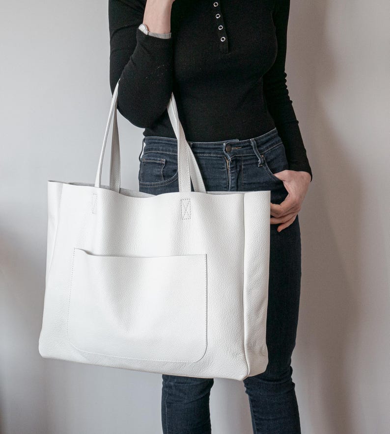 May include: A large, white leather tote bag with a front pocket. The bag has two long handles and is held by a person. The bag is rectangular and appears to be made of a textured leather material. The background is a plain, light gray wall.
