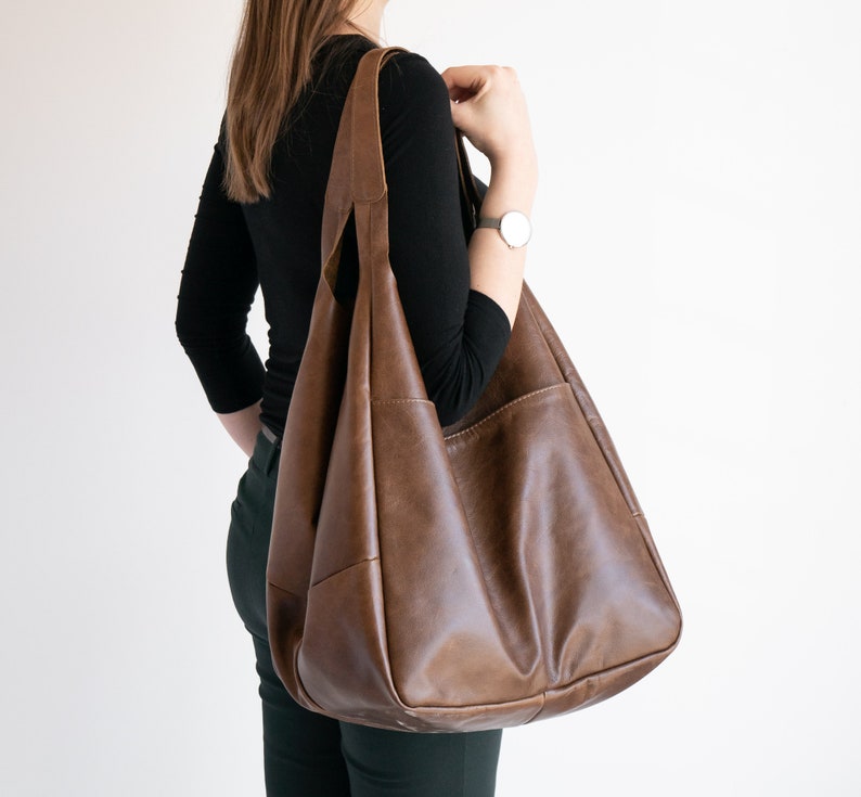 K&ouml;nnte beinhalten: Eine gro&szlig;e braune Ledertasche mit einer breiten &Ouml;ffnung und einem einzigen Trageriemen. Die Tasche hat ein ger&auml;umiges Innenleben und einen l&auml;ssigen, entspannten Stil.