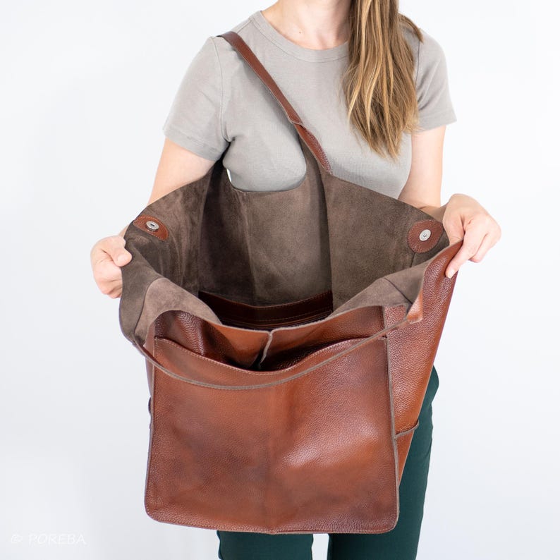 May include: A large brown leather tote bag with a brown suede lining. The bag is open and the lining is visible. The bag has a single strap and a zippered pocket on the front.