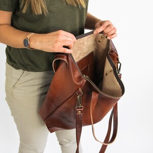 May include: A large brown leather tote bag with a textured exterior, a zipper closure, and an interior pocket. The bag features two leather handles and a detachable shoulder strap with antique brass-toned metal hardware clips.