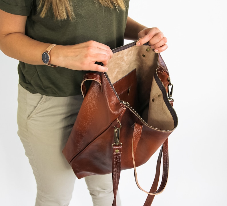 May include: A large brown leather tote bag with a textured exterior, a zipper closure, and an interior pocket. The bag features two leather handles and a detachable shoulder strap with antique brass-toned metal hardware clips.