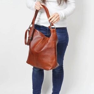 May include: A brown leather hobo bag with a long strap. The bag is being held by a person wearing blue jeans and a white sweater.