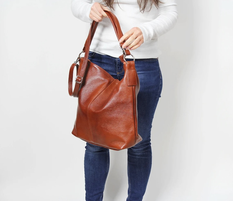 May include: A brown leather hobo bag with a long strap. The bag is being held by a person wearing blue jeans and a white sweater.