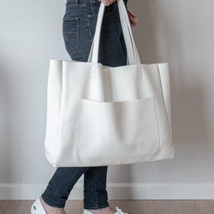 May include: A large, white tote bag with a front pocket and long handles. The bag is held by a person wearing dark blue jeans and white shoes. The bag appears to be made of a textured material.