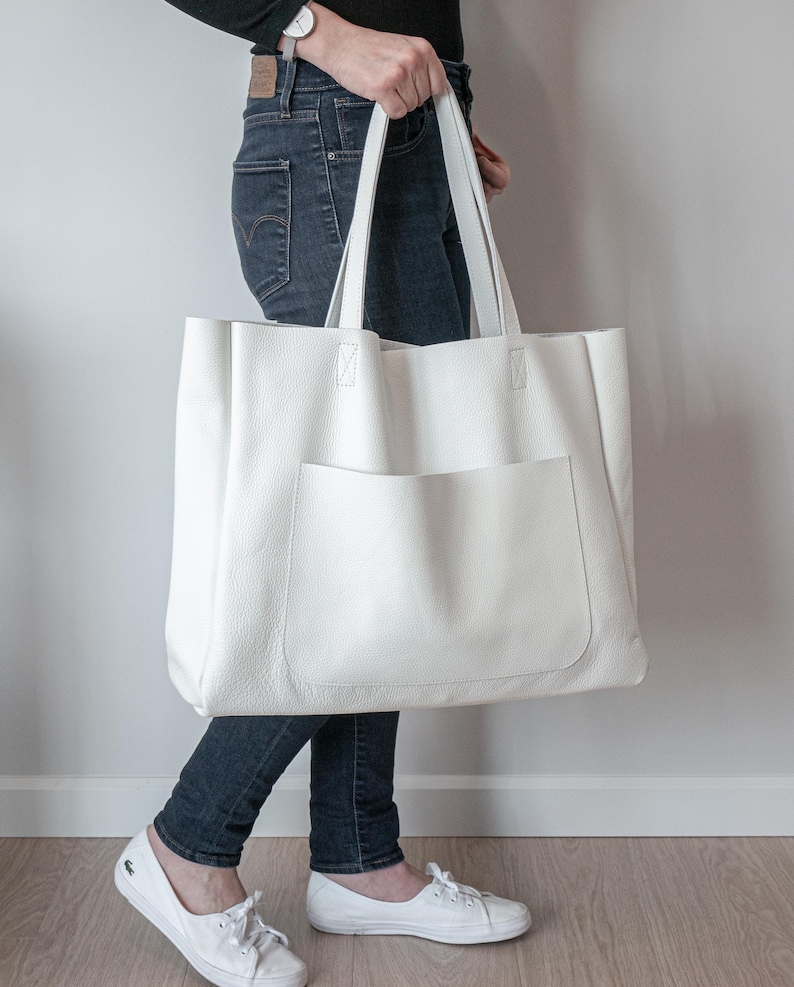 May include: A large, white tote bag with a front pocket and long handles. The bag is held by a person wearing dark blue jeans and white shoes. The bag appears to be made of a textured material.