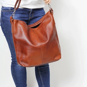 May include: A brown leather tote bag with a long strap. The bag is worn over the shoulder of a person wearing blue jeans.
