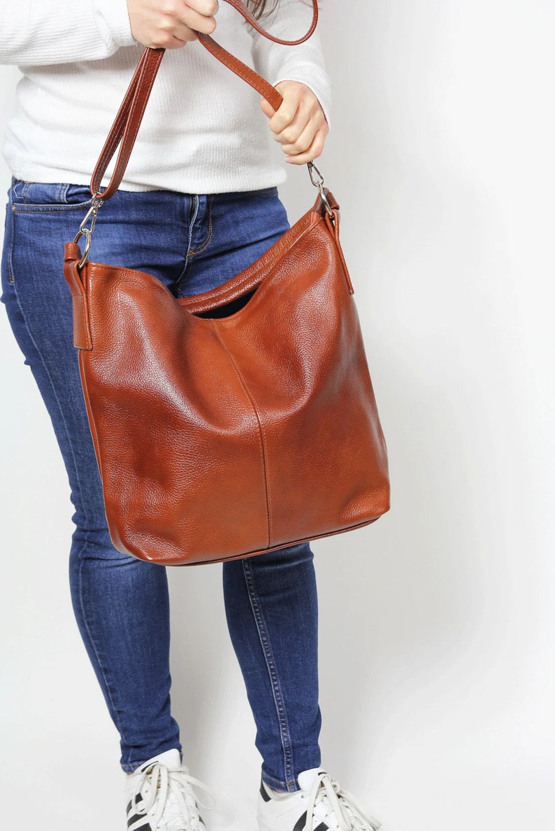 May include: A brown leather tote bag with a long strap. The bag is worn over the shoulder of a person wearing blue jeans.