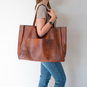 May include: A large brown leather tote bag with a front pocket. The bag is being held by a person wearing a light brown shirt and blue jeans.