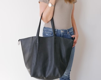 May include: A large black tote bag with a pebbled texture, featuring two long shoulder straps and a silver-toned metal ring detail on the side. The bag has a wide, open-top design with a slouchy silhouette and vertical seam detailing.