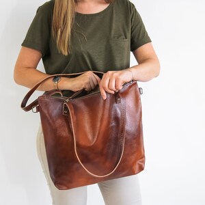 May include: A large, rectangular brown leather tote bag with a smooth, polished finish. The bag features two thin, light-colored leather shoulder straps, a top zipper closure, and metal hardware attachments on the sides for an optional strap.