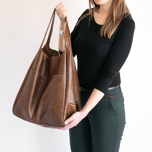 May include: A large, slouchy brown leather tote bag featuring two long shoulder straps and a spacious main compartment. The exterior surface has a smooth, slightly distressed finish with visible stitching details along the side pockets.
