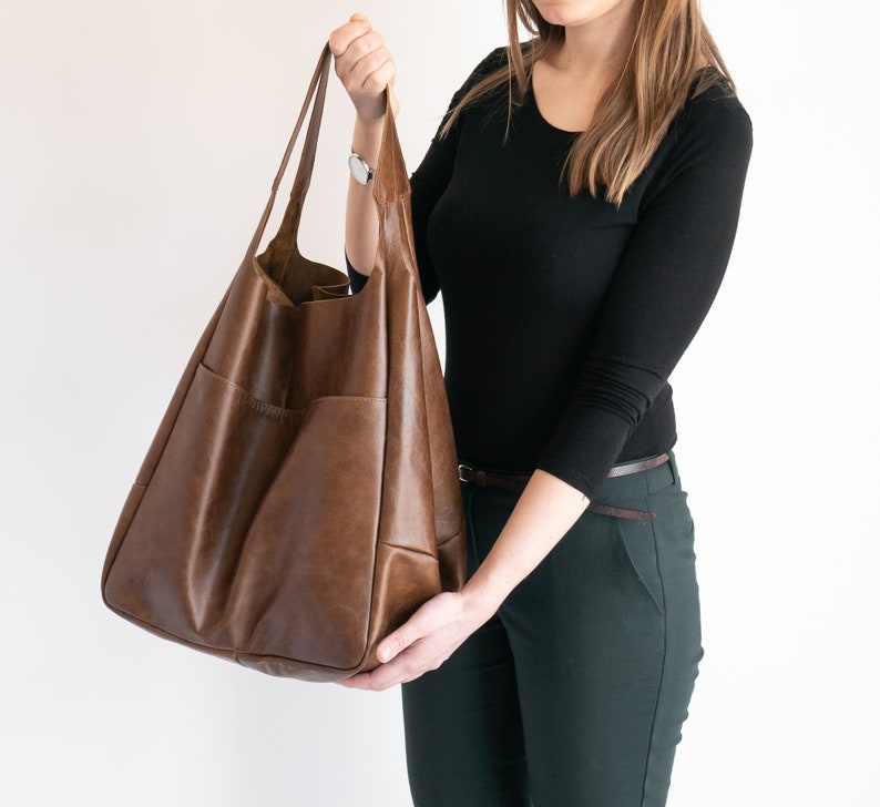 May include: A large, slouchy brown leather tote bag featuring two long shoulder straps and a spacious main compartment. The exterior surface has a smooth, slightly distressed finish with visible stitching details along the side pockets.