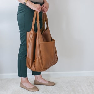 K&ouml;nnte beinhalten: Eine gro&szlig;e, weiche Tragetasche aus hellbraunem Leder mit zwei langen Schulterriemen und einer auff&auml;lligen Au&szlig;entasche auf der Vorderseite. Die Tasche hat eine lockere Form mit sichtbaren N&auml;hten an der Unterseite und den Seiten.