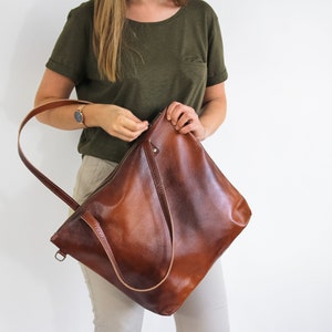 May include: A large, structured brown leather tote bag with a smooth, polished finish. The bag features a single long shoulder strap, a top zipper closure, and a small metal D-ring attachment on the side for added utility and design.