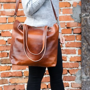 May include: A large, cognac-brown leather tote bag with a smooth, slightly pebbled texture. It features two sturdy top handles, a long adjustable shoulder strap with metal hardware, and a horizontal zipper closure across the top opening.