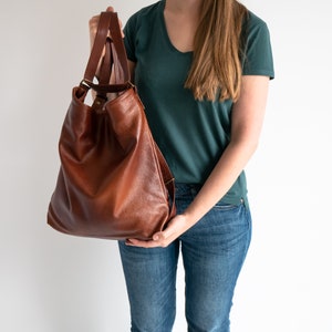 Peut inclure: Un sac fourre-tout en cuir marron avec une longue bandoulière. Le sac est tenu par une personne portant un t-shirt turquoise et un jean bleu.