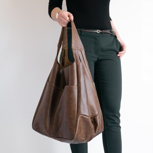 May include: A large, slouchy brown hobo bag featuring a smooth, matte finish, two integrated shoulder straps, and a spacious main compartment with visible exterior pockets on the front panel, crafted with a soft, flexible structure.