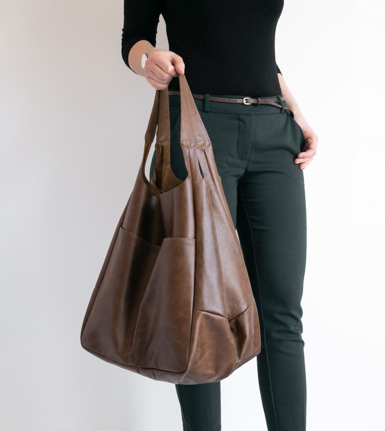 May include: A large, slouchy brown hobo bag featuring a smooth, matte finish, two integrated shoulder straps, and a spacious main compartment with visible exterior pockets on the front panel, crafted with a soft, flexible structure.