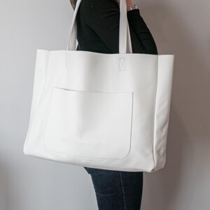 May include: A large, white tote bag with a rectangular front pocket. The bag has two sturdy handles and is made of a smooth, solid material. The bag is being held by a person wearing a black long-sleeve shirt and blue jeans.