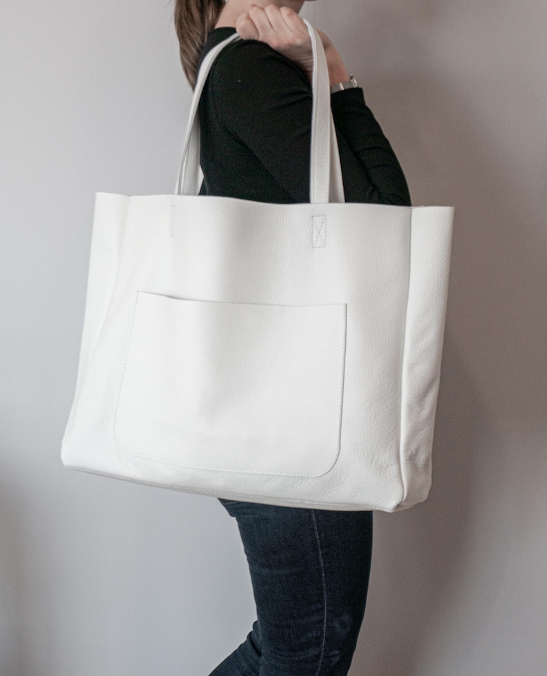 May include: A large, white tote bag with a rectangular front pocket. The bag has two sturdy handles and is made of a smooth, solid material. The bag is being held by a person wearing a black long-sleeve shirt and blue jeans.