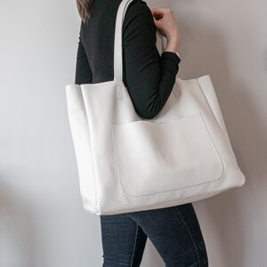 May include: A large, white tote bag with a textured surface and a front pocket. The bag has two long straps for carrying over the shoulder. The bag is made of a soft material and is a neutral color.