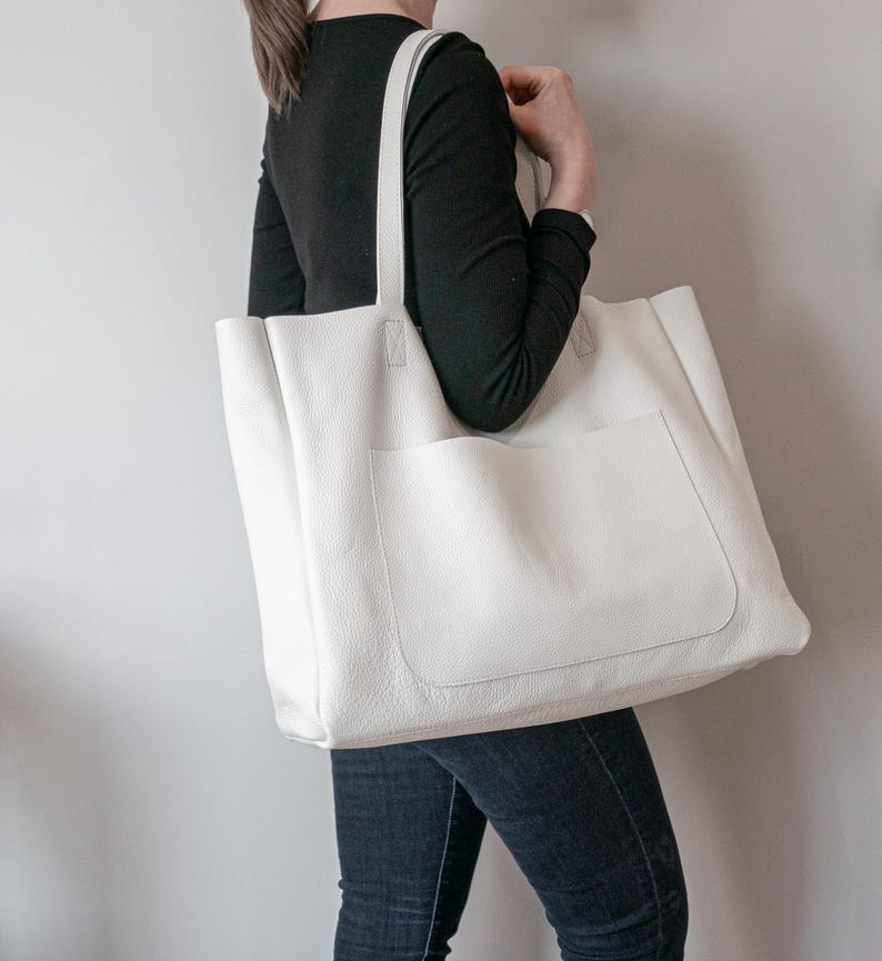 May include: A large, white tote bag with a textured surface and a front pocket. The bag has two long straps for carrying over the shoulder. The bag is made of a soft material and is a neutral color.