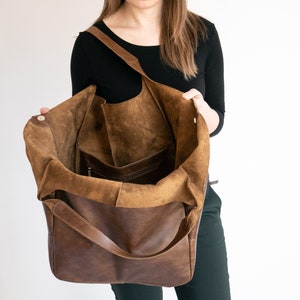 May include: A large brown leather hobo bag with a wide shoulder strap and an open top. The interior features a soft, unlined suede texture and includes a single internal pocket with a zipper closure for secure storage of small items.