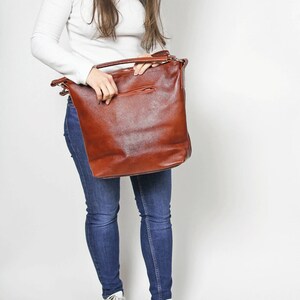May include: A brown leather tote bag with a zippered pocket on the front. The bag is being held by a person wearing a white shirt and blue jeans.