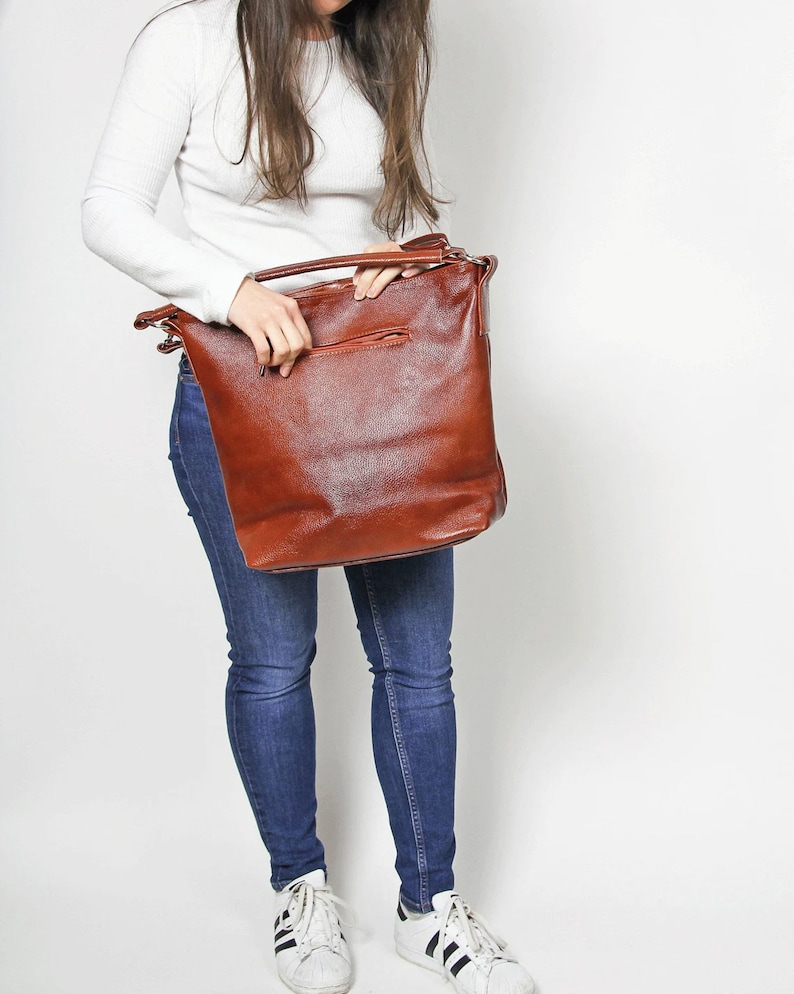 May include: A brown leather tote bag with a zippered pocket on the front. The bag is being held by a person wearing a white shirt and blue jeans.