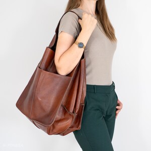 May include: A brown leather tote bag with a large pocket on the front. The bag is being carried by a person wearing a light brown t-shirt and dark green pants.