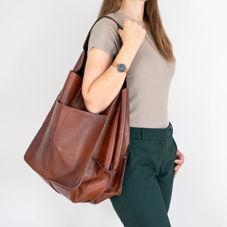 May include: A brown leather tote bag with a large pocket on the front. The bag is being carried by a person wearing a light brown t-shirt and dark green pants.