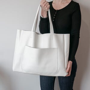 May include: A large, white leather tote bag with a front pocket. The bag has two long handles and is held by a person. The tote bag is a simple, minimalist design, suitable for everyday use. The bag's dimensions are approximately 16 inches wide and 14 inches tall.